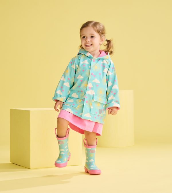 An aqua blue hooded raincoat for younger girls by Hatley, printed with a lovely sky design, featuring sunshine, pink hearts and clouds. Practical to wear on rainy days, it has a smooth waterproof coating and light reflective logo patches with a soft pink terry towelling lining for extra comfort. It fastens with pink poppers on the front.